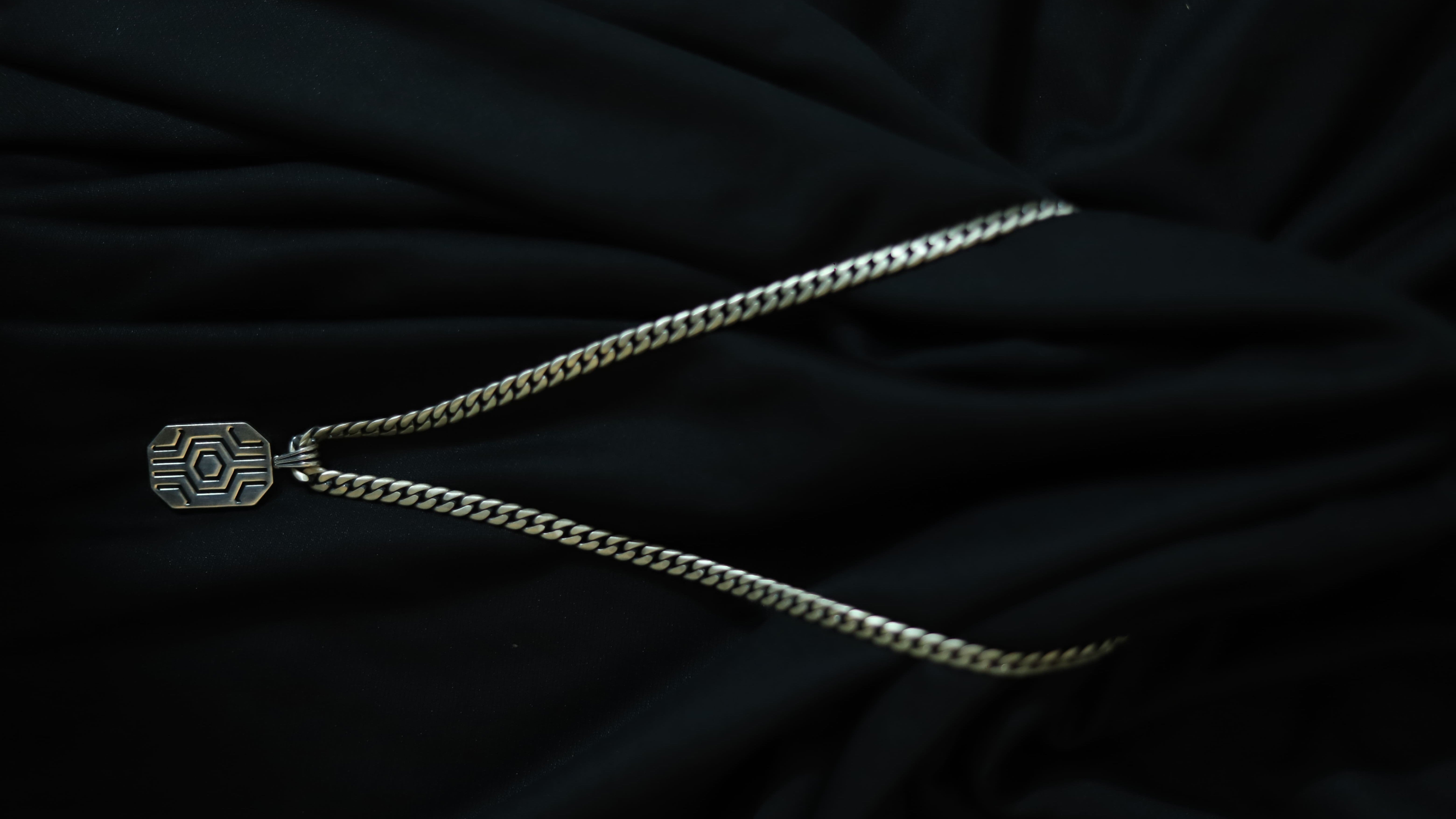 A silver-toned chain necklace with a geometric, hexagonal pendant featuring a sunburst pattern, displayed on a draped black fabric background.